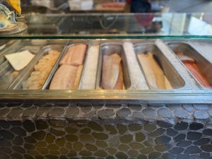 Assorted fish fillets arranged in metal trays behind a glass display at a seafood counter.