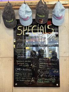 Chalkboard restaurant specials sign with colorful handwriting, hats hung above the menu display, reflecting a casual dining vibe.