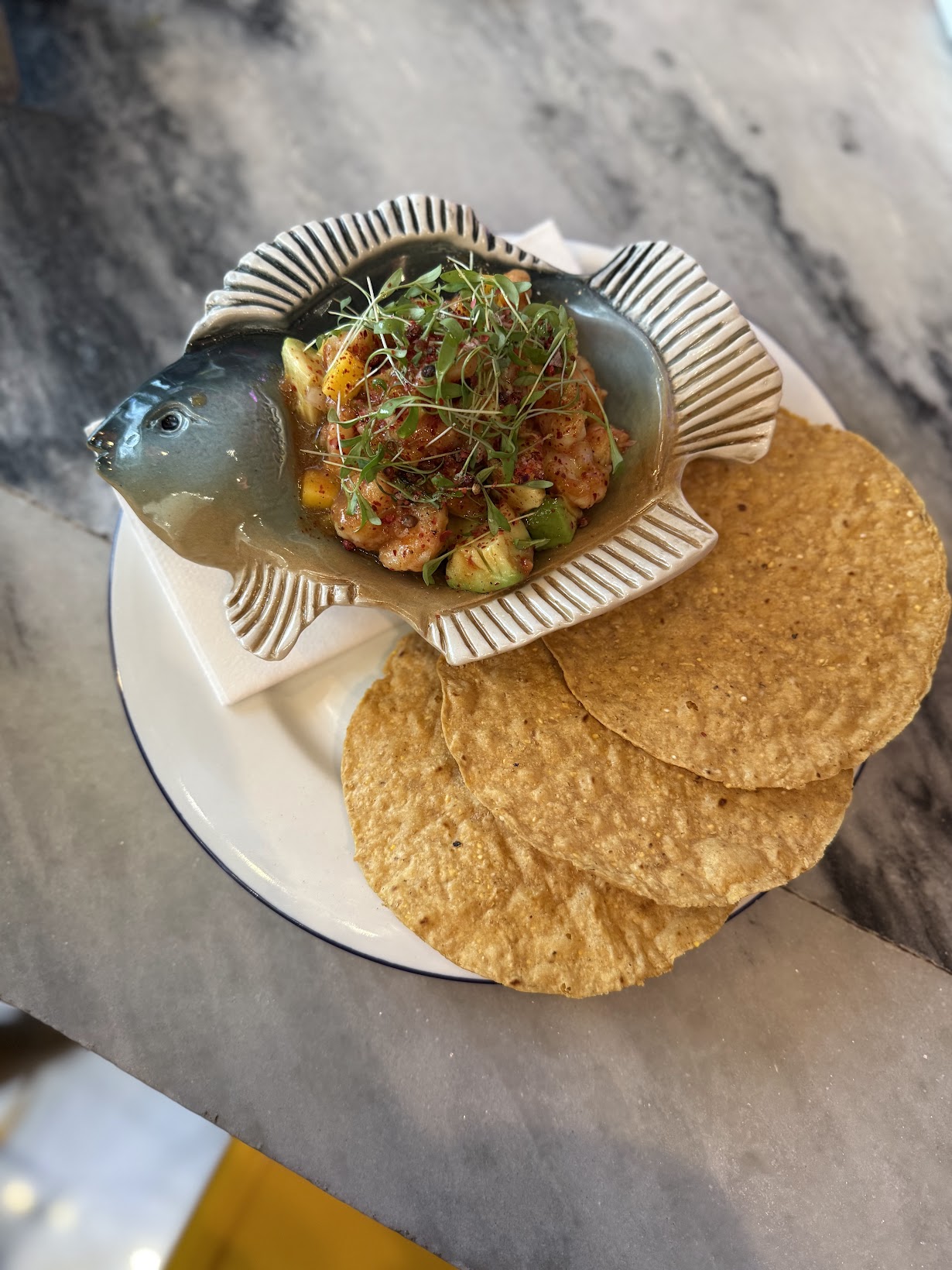 Fish-shaped ceramic bowl holding a shrimp and veggie salsa topped with microgreens, served with several corn tortillas on a white plate.