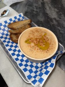 Creamy bowl of soup with olive oil drizzle and paprika, served with toasted bread on a checkered liner.
