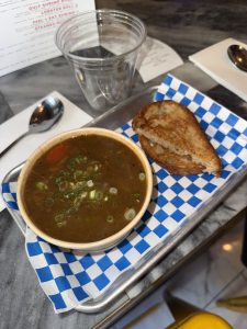 Bowl of dark broth soup garnished with chopped green onions, with a slice of toasted bread on a metal tray lined by blue-and-white checkered paper.