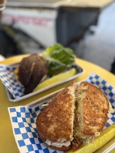 Half a crusty sandwich filled with meat and veggies on a yellow plate, with pickles nearby and blue-checkered paper underneath.