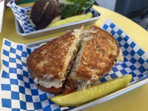 Grilled sandwich halves with melted cheese and sprouts served on checkered paper in a metal tray with a pickle spear nearby.